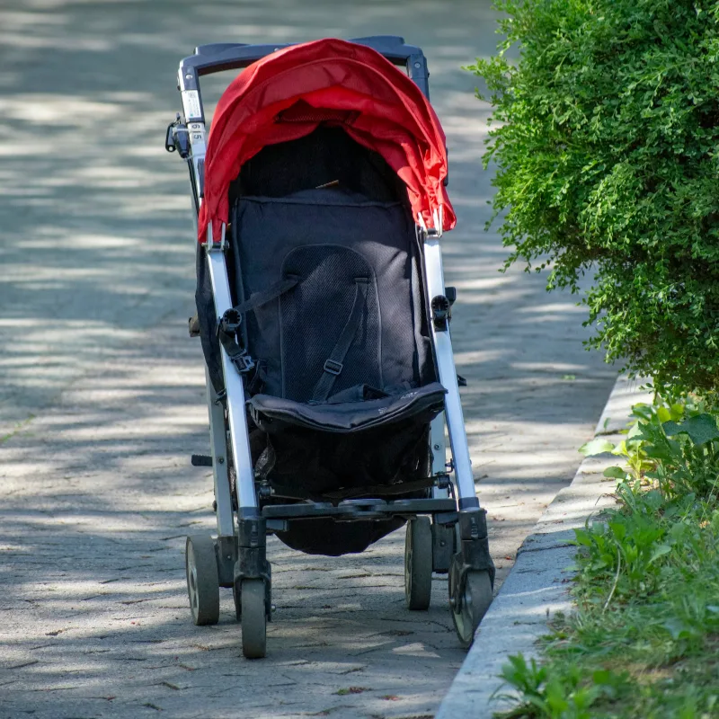 Carrinho de bebê vermelho e preto, vazio, numa calçada ao lado de arbustos
