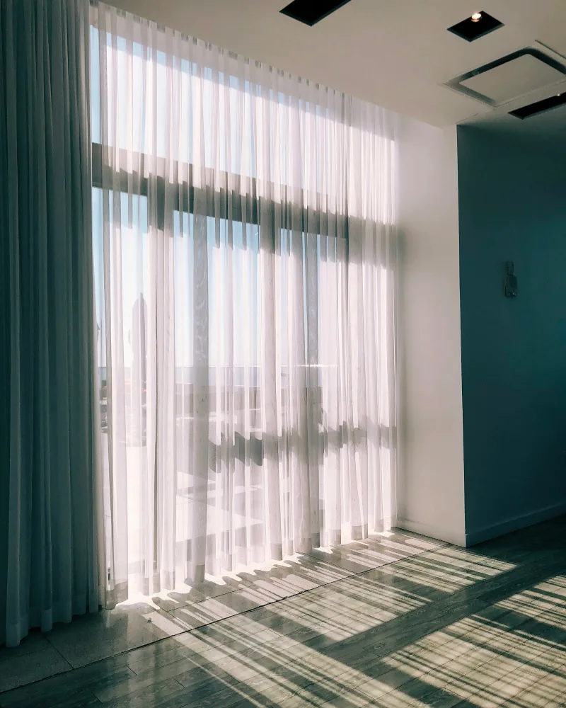 Cortinas brancas transparentes numa janela grande, com sol entrando na sala vazia