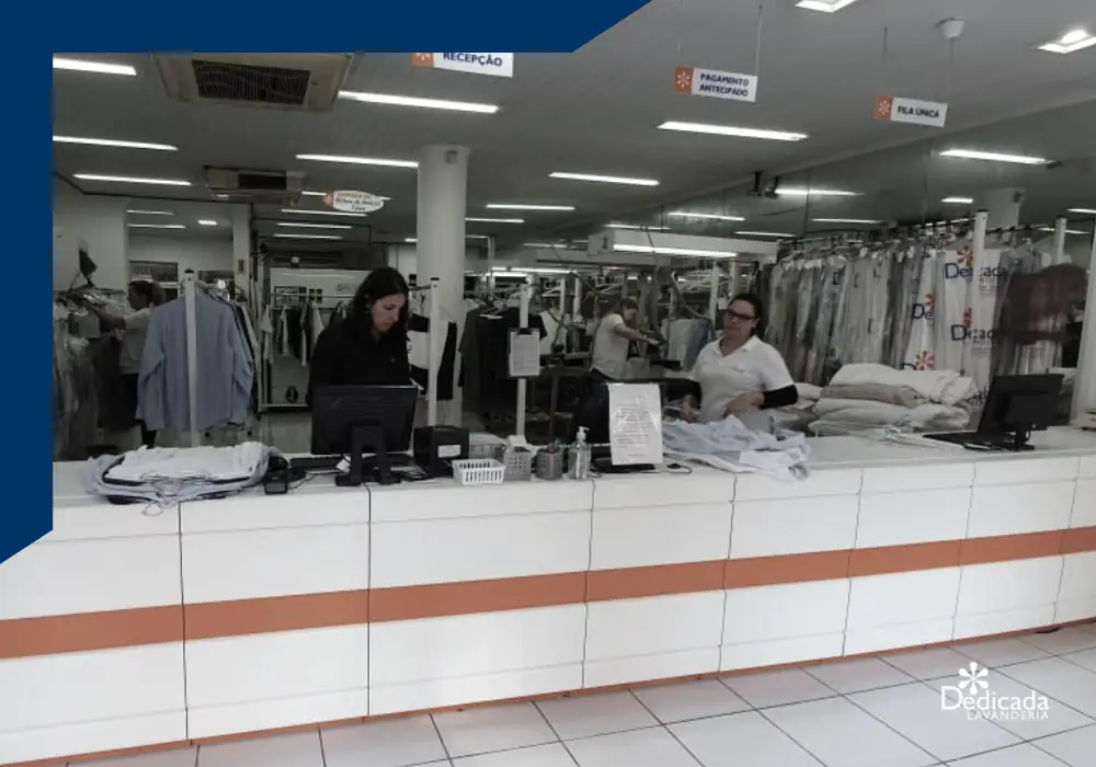 Balcão de atendimento da Dedicada Lavanderia, com duas atendentes conferindo roupas e peças prontas penduradas ao fundo