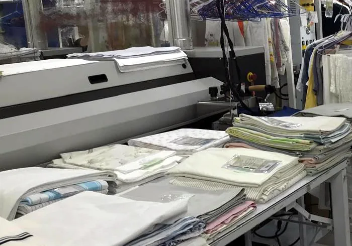Lençóis e fronhas dobrados sobre a mesa ao lado da calandra, na produção da Dedicada Lavanderia