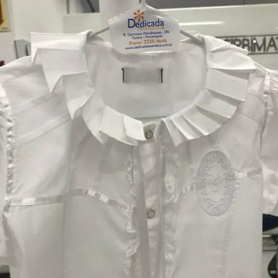 Camisa infantil branca com gola plissada, pronta no cabide com a etiqueta da Dedicada Lavanderia