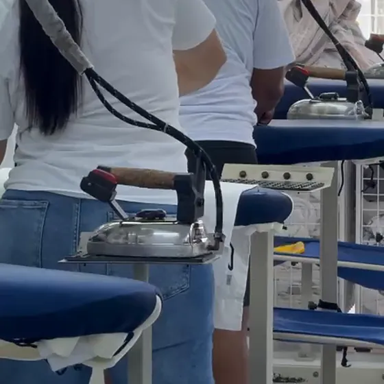 Ferro a vapor no apoio ao lado das mesas de passar, com a equipe de passadoria de costas, na Dedicada Lavanderia