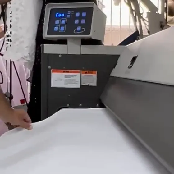 Lençol branco liso guiado à mão na entrada da calandra, com o painel da máquina ao fundo, na Dedicada Lavanderia