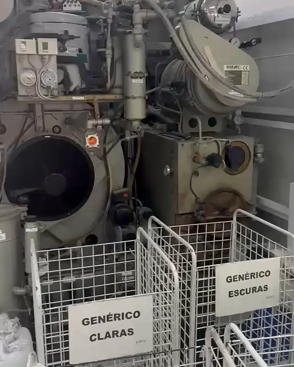 Parte de trás da máquina de lavagem a seco Firbimatic, com dois cestos de arame com as placas "Genérico claras" e "Genérico escuras", na produção da Dedicada Lavanderia