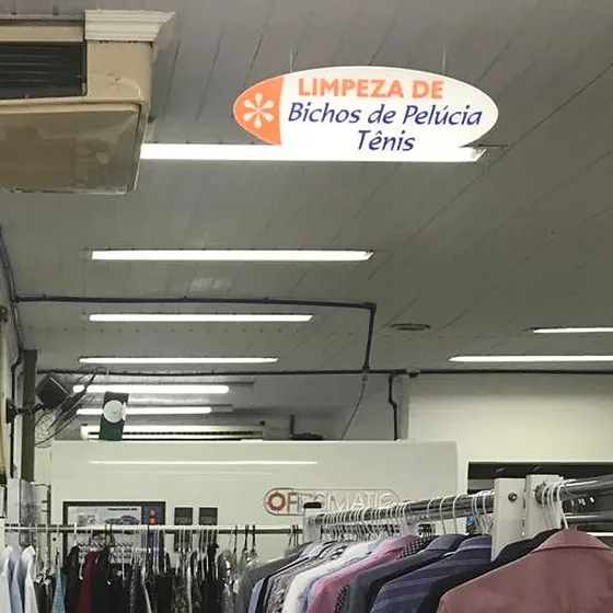 Placa de limpeza de bichos de pelúcia e tênis pendurada no teto do salão da Dedicada Lavanderia, sobre araras de camisas