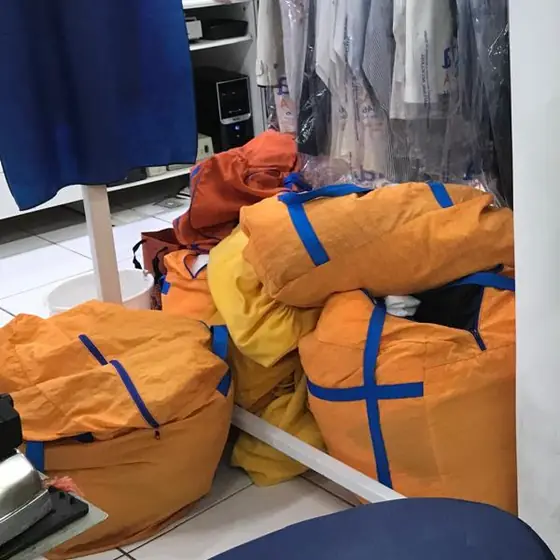 Sacos laranja com roupas recebidas, no chão da produção da Dedicada Lavanderia, ao lado das araras de peças em sacos plásticos