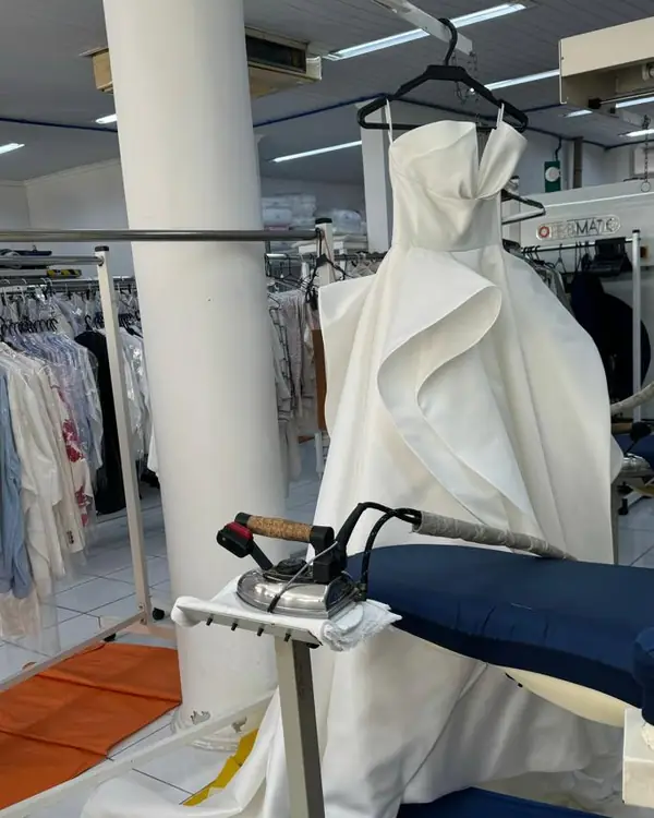 Vestido branco longo, com babado em cascata, no cabide ao lado da mesa de passar com ferro a vapor, na Dedicada Lavanderia
