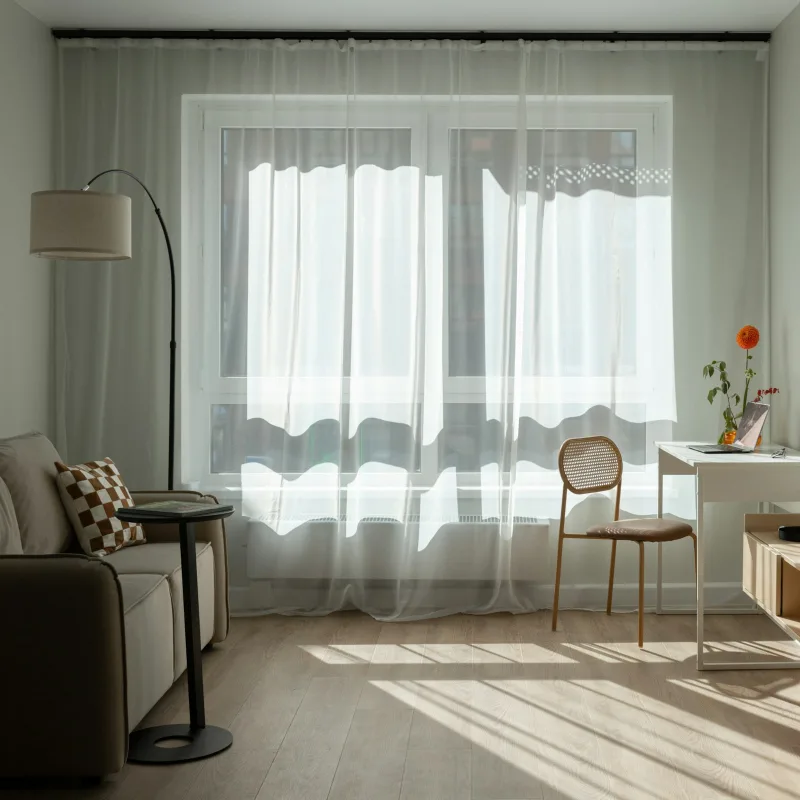 Sala com cortinas brancas transparentes na janela, poltrona e mesa, com luz do dia