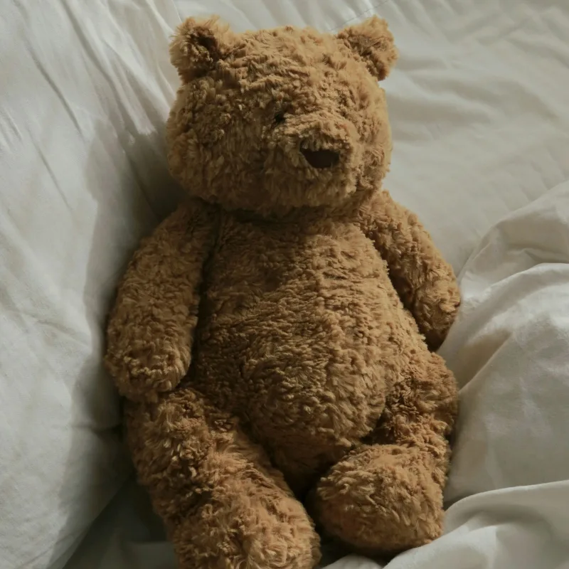 Urso de pelúcia marrom sentado numa cama com lençóis brancos